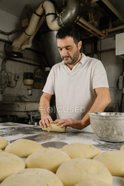 Maduro panadero étnico barbudo formando pan de masa en la mesa con harina y tazón en la panadería - foto de stock