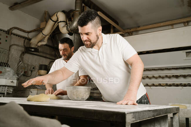 From below mature bearded ethnic bakers forming bread from dough at table with flour and bowl in bakery — Stock Photo