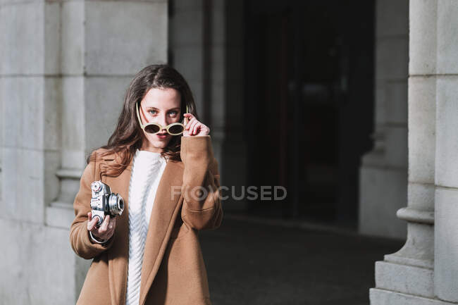 Stylische Frau in Mantel und Sonnenbrille steht mit Vintage-Fotokamera auf der Straße und schaut in die Kamera — Stockfoto