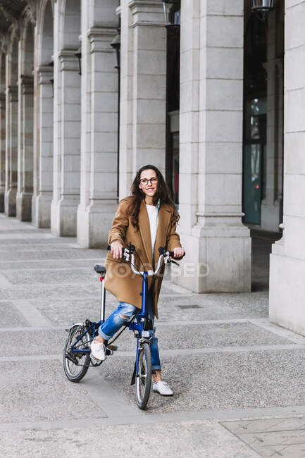 Веселая женщина в пальто смотрит на велосипед против старого здания с колоннами в городе — стоковое фото