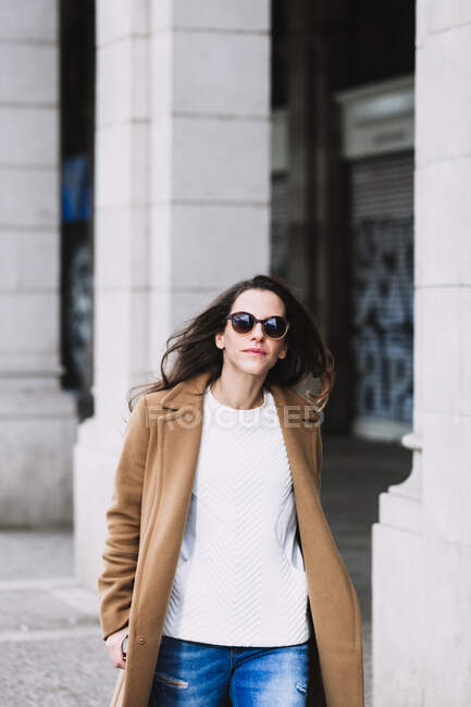 Confident female in coat and modern sunglasses strolling on walkway in city in daylight — Stock Photo