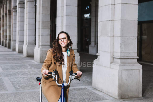Весела жінка в пальто дивиться на велосипед проти старої будівлі з колонами в місті — стокове фото