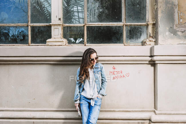 Young female in trendy denim outfit and sunglasses standing with hand in pocket near shabby building in city — Stock Photo