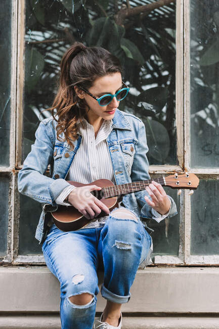Stylish female in denim clothes sitting on windowsill of shabby building and playing ukulele while entertaining at weekend in city — Stock Photo