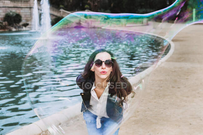 Femme insouciante dans des vêtements à la mode et des lunettes de soleil debout près de la fontaine dans le parc et souffler la bulle de savon — Photo de stock