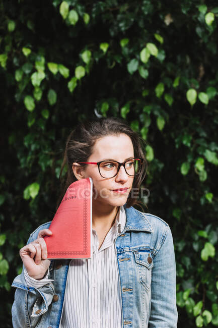 Estudiante femenina contenta en gafas y con bloc de notas de pie en el parque y mirando hacia otro lado - foto de stock