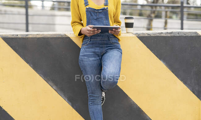 Cultivar fêmea étnica irreconhecível em macacões jeans em pé na rua urbana e assistir a vídeo em tablet na cidade — Fotografia de Stock