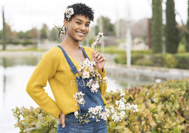 Alegre mujer afroamericana de pie con ramas con flores sobre todo el cuerpo en el parque en primavera y mirando a la cámara - foto de stock