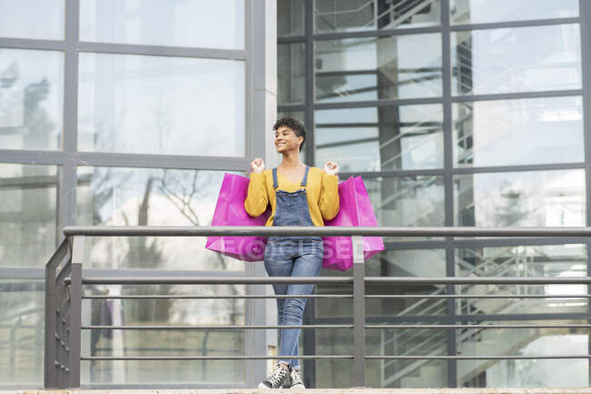 Encantada hembra afroamericana de pie con bolsas de papel en la ciudad y mirando hacia otro lado después de compras exitosas - foto de stock