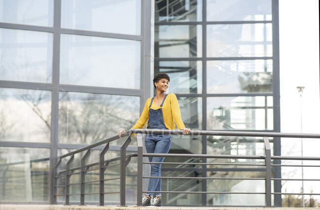 Cheerful African American female in trendy overalls and sneakers standing near building with glass walls in city and looking away — Stock Photo