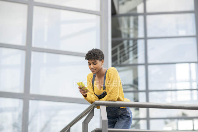 Low angle of content African American female standing near glass building in city and surfing Internet on smartphone — Stock Photo