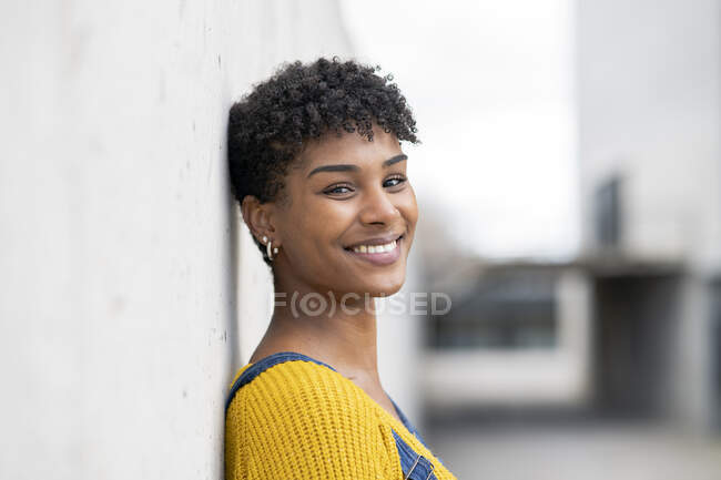 Vista lateral de una mujer afroamericana positiva en overol denim y con peinado afro apoyado en la pared y mirando a la cámara - foto de stock
