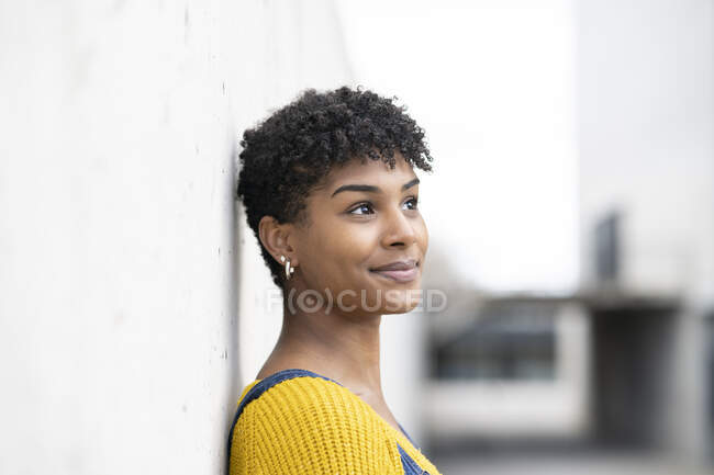 Seitenansicht einer positiven Afroamerikanerin in Jeans-Overalls und mit Afro-Frisur, die sich an die Wand lehnt und wegschaut — Stockfoto