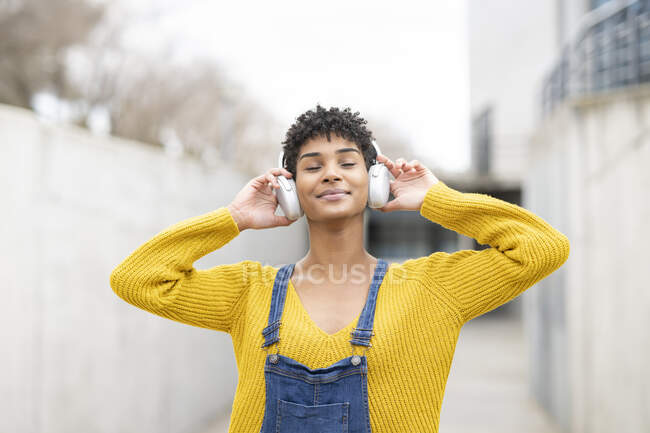 Sonhador sorrindo afro-americano fêmea em fones de ouvido sem fio desfrutando de músicas com olhos fechados enquanto estava na rua — Fotografia de Stock