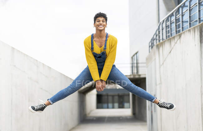Vista lateral da ativa alegre afro-americana vestindo macacões e tênis no momento de saltar acima da rua da cidade — Fotografia de Stock