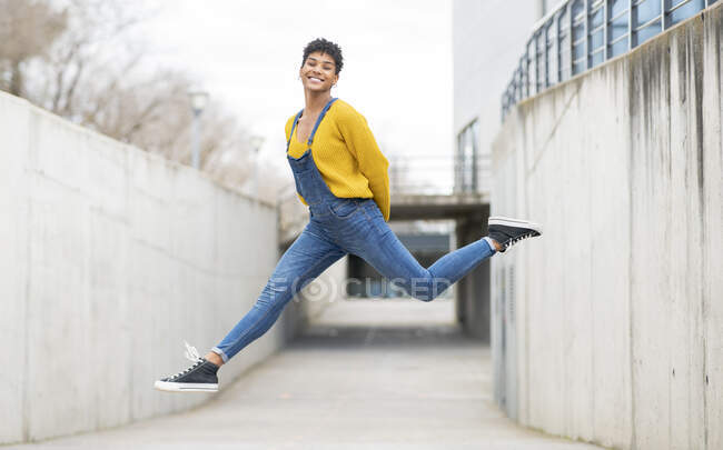 Бічний вид веселої афроамериканської жінки, одягненої в комбінезон і кросівки в момент стрибка над міською вулицею — стокове фото