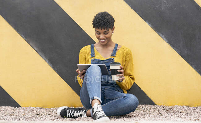 Fêmea afro-americana legal sentada com bebida takeaway em copo de papel perto da parede na cidade e navegar na Internet em tablet — Fotografia de Stock