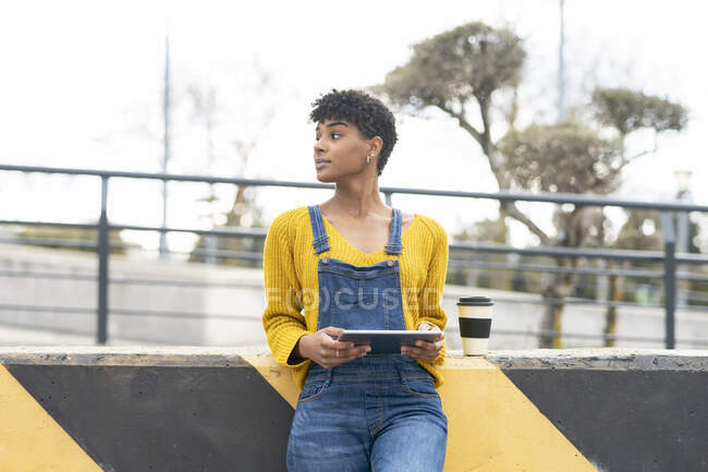 Cool hembra afroamericana con bebida para llevar en taza de papel cerca de la pared en la ciudad y navegar por Internet en la tableta - foto de stock