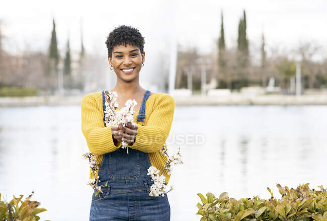 Joyful African American female standing with branches with flowers in park in spring and looking at camera — Stock Photo