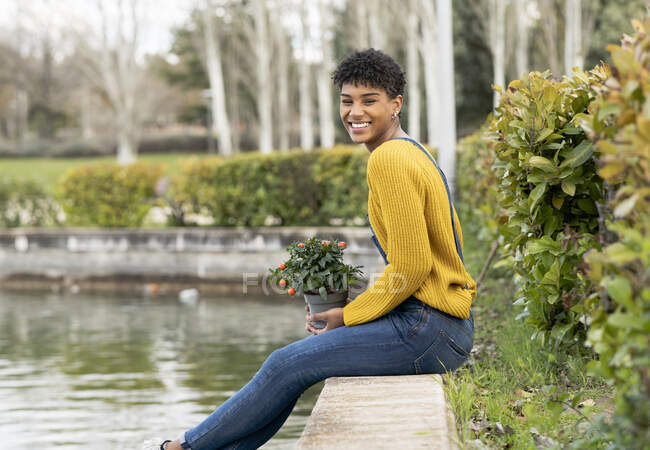 Seitenansicht einer entzückten schwarzen Frau, die in der Nähe eines Teiches im Park mit Topfpflanze sitzt und in die Kamera blickt — Stockfoto