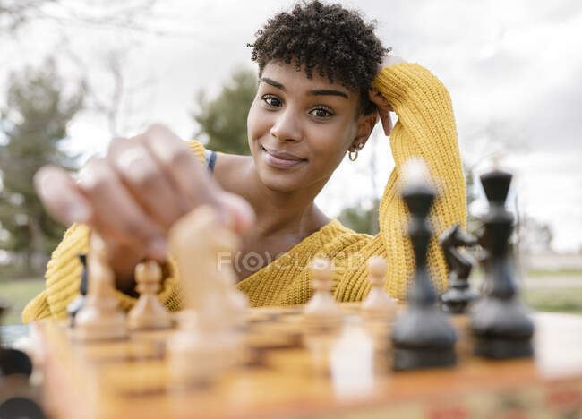 Von unten eine optimistische Afroamerikanerin, die im Park am Tisch sitzt und Schach spielt, während sie in die Kamera schaut — Stockfoto