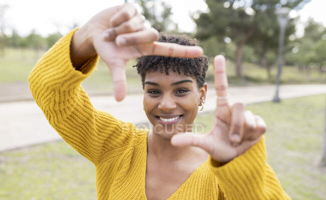 Позитивная афроамериканка с вьющимися волосами, стоящая в парке и показывающая вывеску в рамке, глядя в камеру — стоковое фото