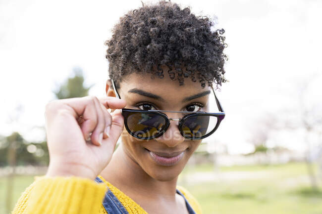 Femme afro-américaine souriante aux cheveux bouclés touchant des lunettes de soleil à la mode et regardant la caméra dans le parc — Photo de stock