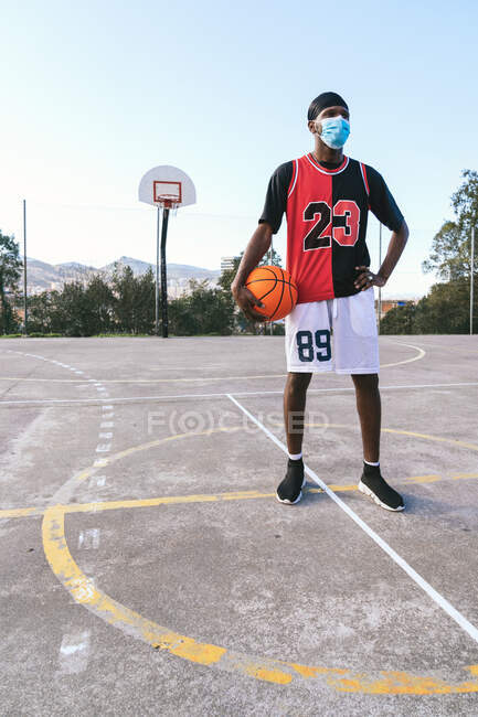 Jogador de streetball masculino afro-americano determinado em máscara e com bola em pé no playground de basquete e olhando para longe — Fotografia de Stock