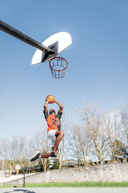 Dal basso del giocatore di streetball afroamericano maschile nel momento di saltare e segnare pallacanestro nel cerchio — Foto stock
