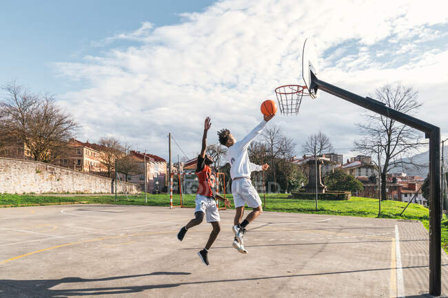 Vista lateral de un hombre afroamericano y un amigo hispano jugando streetball mientras salta y anota en el aro - foto de stock