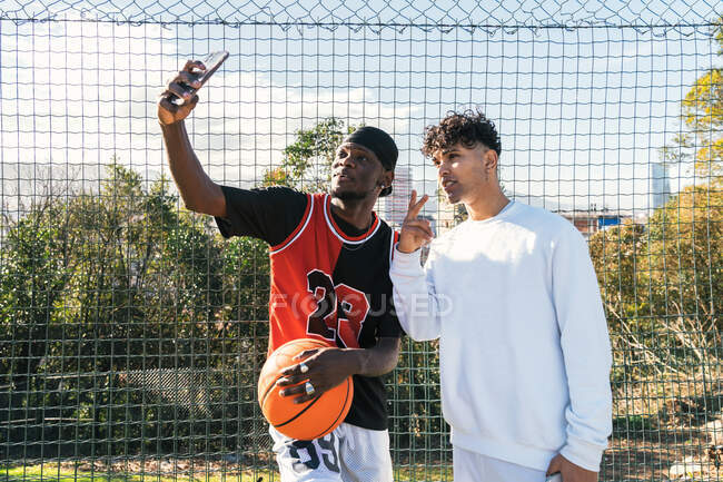 Young African American male streetball players standing on basketball playground and taking selfie on smartphone — Stock Photo