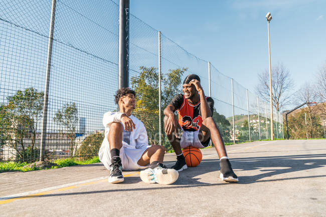 Sorridenti giocatori di streetball afroamericani maschili seduti sul campo da basket nella giornata di sole e guardarsi l'un l'altro — Foto stock