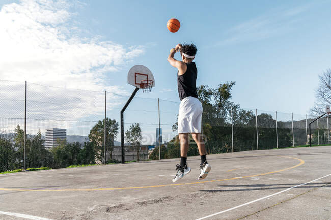 Vista lateral del jugador de streetball masculino étnico realizando slam dunk en el momento de saltar por encima del patio de recreo y anotar baloncesto en el aro - foto de stock