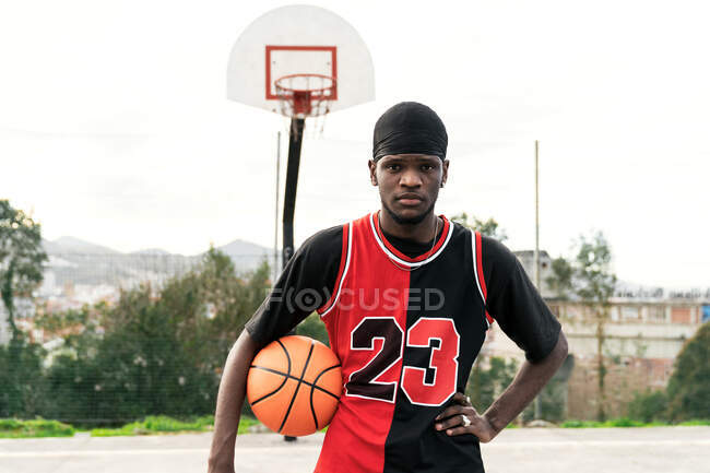 Sério afro-americano jogador de streetball masculino em uniforme de pé com bola na quadra de basquete e olhando para a câmera — Fotografia de Stock