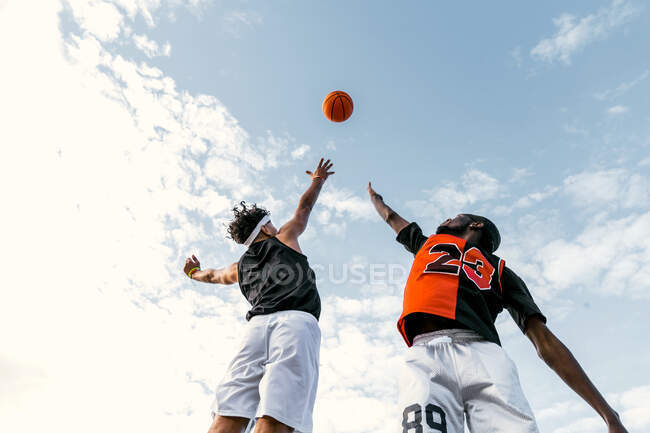 From below of African American male friends playing streetball and jumping on background of blue sky in summer — Stock Photo