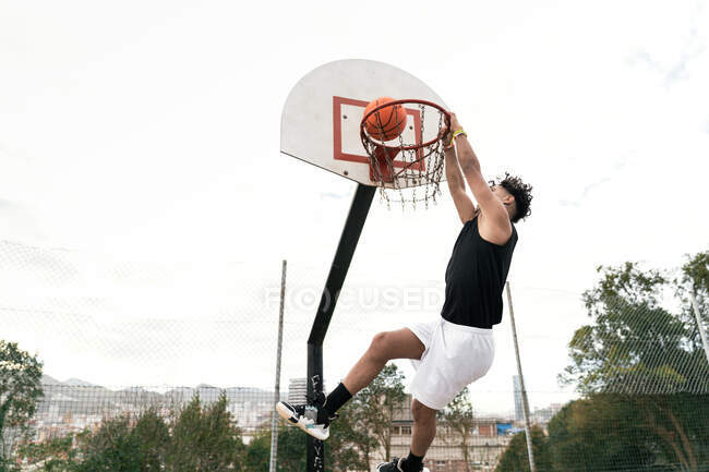 Vista lateral do jogador de streetball masculino étnico realizando slam dunk no momento de saltar acima do playground e marcar basquete em aro — Fotografia de Stock