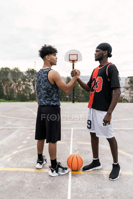 Side view of African American male streetball players shaking hands while standing on basketball playground and looking at each other — Stock Photo