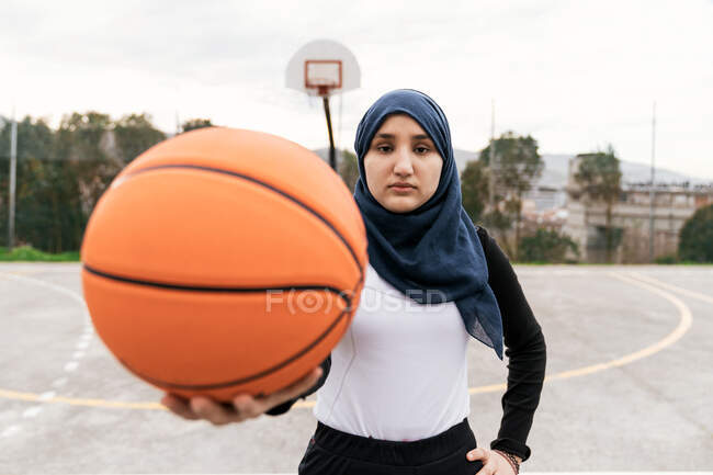 Joueuse de streetball musulmane confiante dans le hijab debout avec une balle sur le terrain de basket et regardant la caméra — Photo de stock