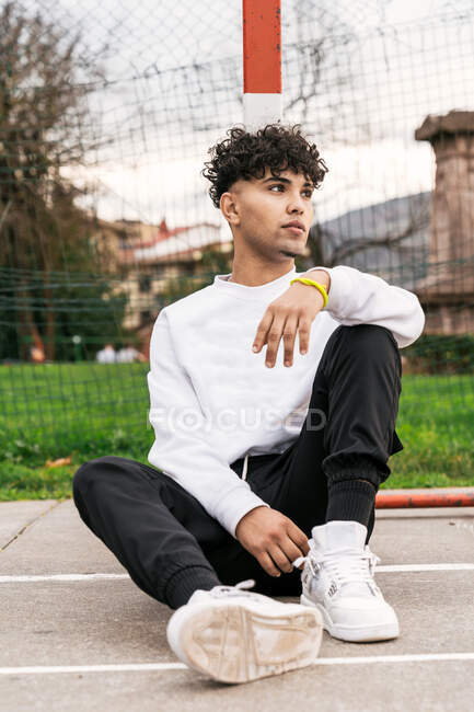 Young African American male in trendy clothes sitting on basketball playground and looking away — Stock Photo