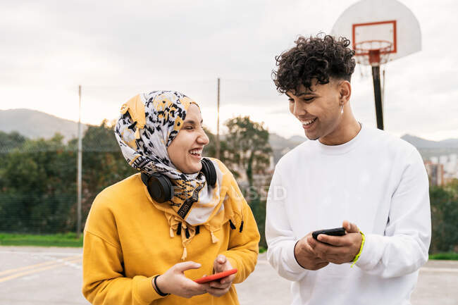Delighted diverse friends standing on basketball playground and browsing smartphones while laughing and having fun in city — Stock Photo