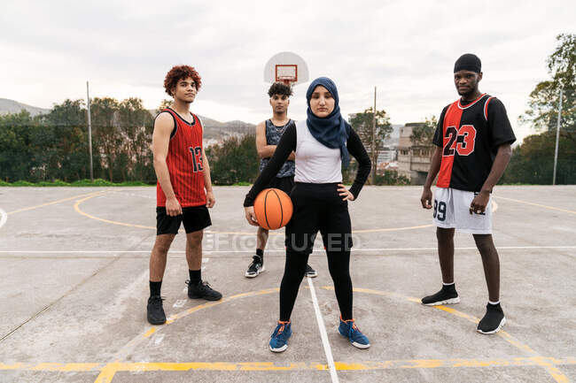 Selbstbewusste multiethnische Streetballmannschaft mit Ball steht auf Basketballsportplatz in der Stadt und blickt auf cmaera — Stockfoto