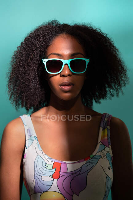 Atractiva mujer afroamericana con traje de baño de moda en gafas de sol mientras está de pie sobre fondo azul y mirando a la cámara en la sombra media - foto de stock