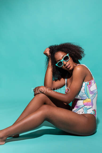 Full body feminine cheerful African American female wearing stylish swimsuit touching curly hair and sitting with sunglasses on blue studio background — Stock Photo