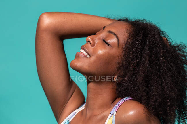 Side view of feminine cheerful African American female wearing stylish swimsuit touching curly hair with eyes closed on blue studio background — Stock Photo