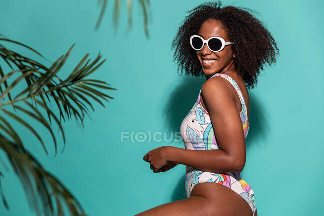Vista lateral da mulher afro-americana atraente feliz vestindo maiô colorido e óculos de sol e olhando para a câmera com sorriso enquanto sentado em fundo azul — Fotografia de Stock