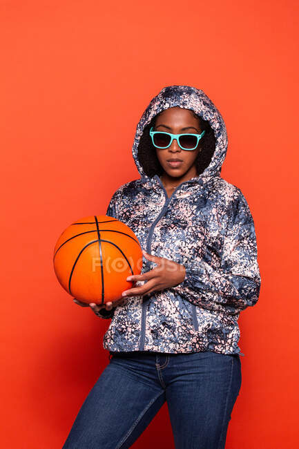 Модная афроамериканка в уличной одежде и прохладных солнцезащитных очках стоит с баскетбольным мячом в руках на красном фоне — стоковое фото