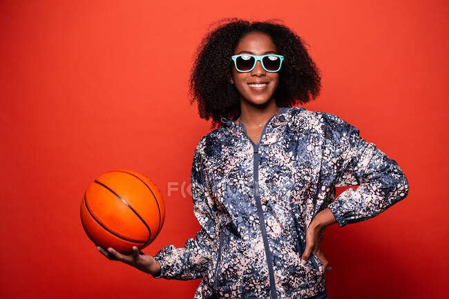 Feminino afro-americano na moda no estilo de rua desgaste e óculos de sol legal de pé com bola de basquete nas mãos contra o fundo vermelho — Fotografia de Stock