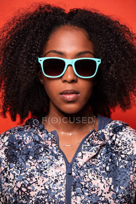 Mulher afro-americana atraente confiante na roupa da moda e óculos de sol azuis olhando para a câmera contra o fundo vermelho — Fotografia de Stock