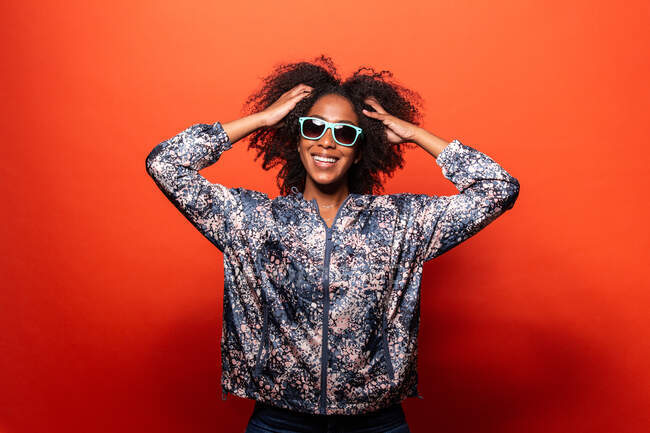 Heureuse jolie femme afro-américaine en tenue tendance et lunettes de soleil bleues regardant la caméra sur fond rouge avec les mains touchant les cheveux — Photo de stock