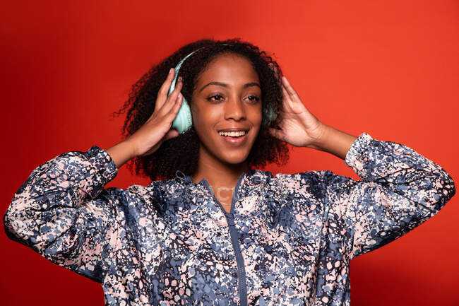 Joven mujer afroamericana alegre en traje elegante escuchando música fresca en auriculares inalámbricos y mirando hacia otro lado con sonrisa contra la pared roja - foto de stock